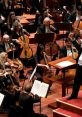 Orchestra performing classical music on stage, conductor leading musicians with string instruments in a concert hall.