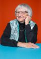 Smiling elderly woman with short gray hair, wearing glasses and a turquoise vest, against a vibrant orange background.