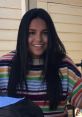 Cheerful young woman with long dark hair in a colorful striped sweater, smiling at a table with notes in a casual setting.