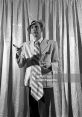 Dapper man in a light suit holding a cigarette, speaking animatedly against a textured backdrop. Classic British style.